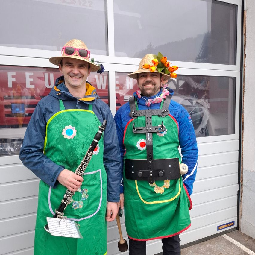 Zwei Männer, gekleidet in grüne Schürzen und Hüte, lächeln für ein Foto vor einem Gebäude mit einem reflektierten Feuerwehrauto. Einer hält eine Klarinette, und der andere hat einen Gürtel mit Rädern.