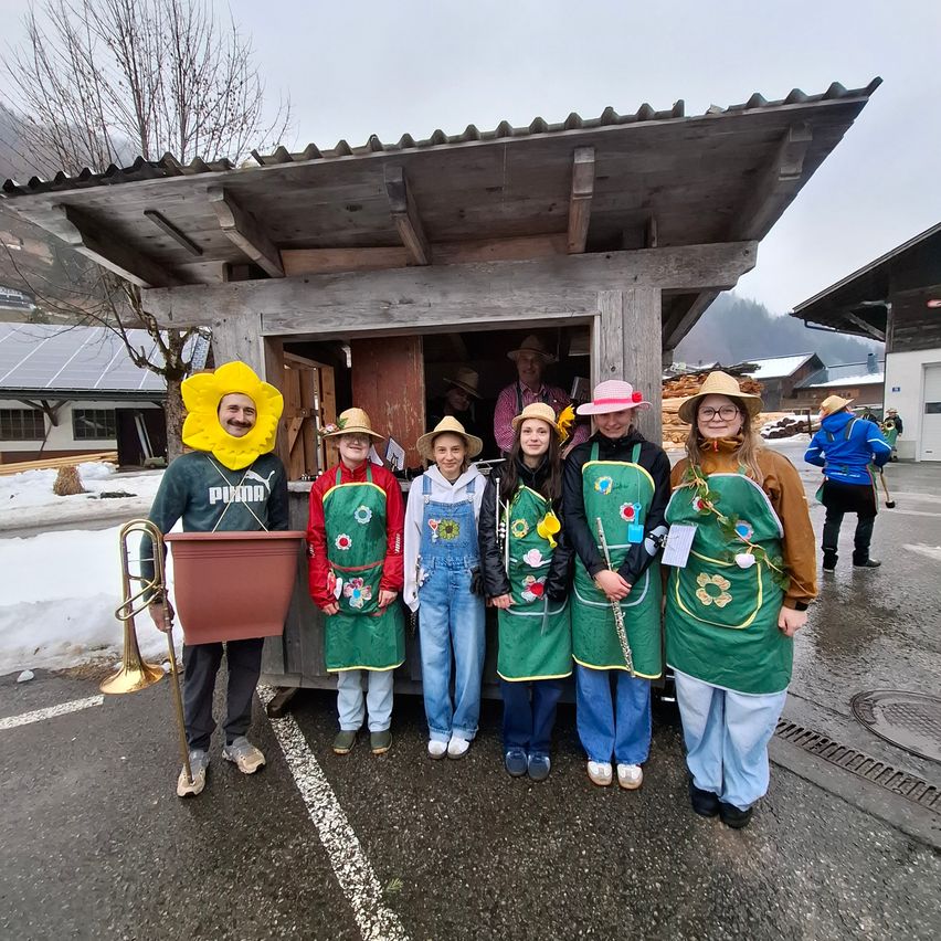 Eine Gruppe von Menschen in Blumenkostümen posiert vor einer Holzhütte, mit Schnee und einem Berg im Hintergrund.