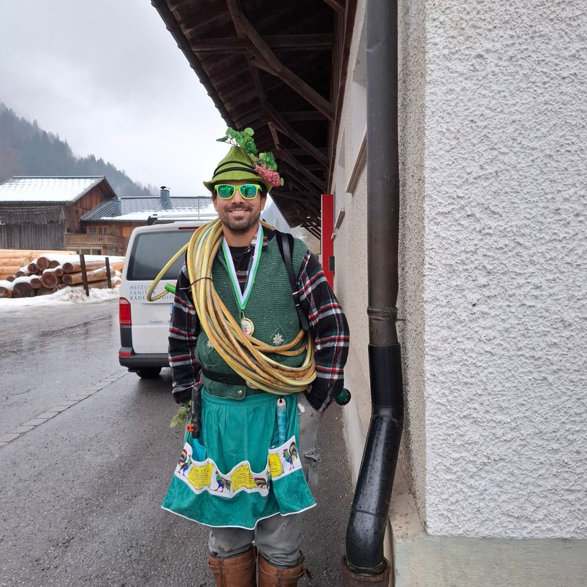 Ein Mann in einem Kostüm steht draußen und lächelt. Er trägt einen grünen Hut mit einer Blume, eine Sonnenbrille und eine grüne Weste mit Medaillen. Eine Schlange und Werkzeuge hängen um ihn herum. Ein Gebäude und ein Van sind hinter ihm, mit Bergen im Hintergrund.