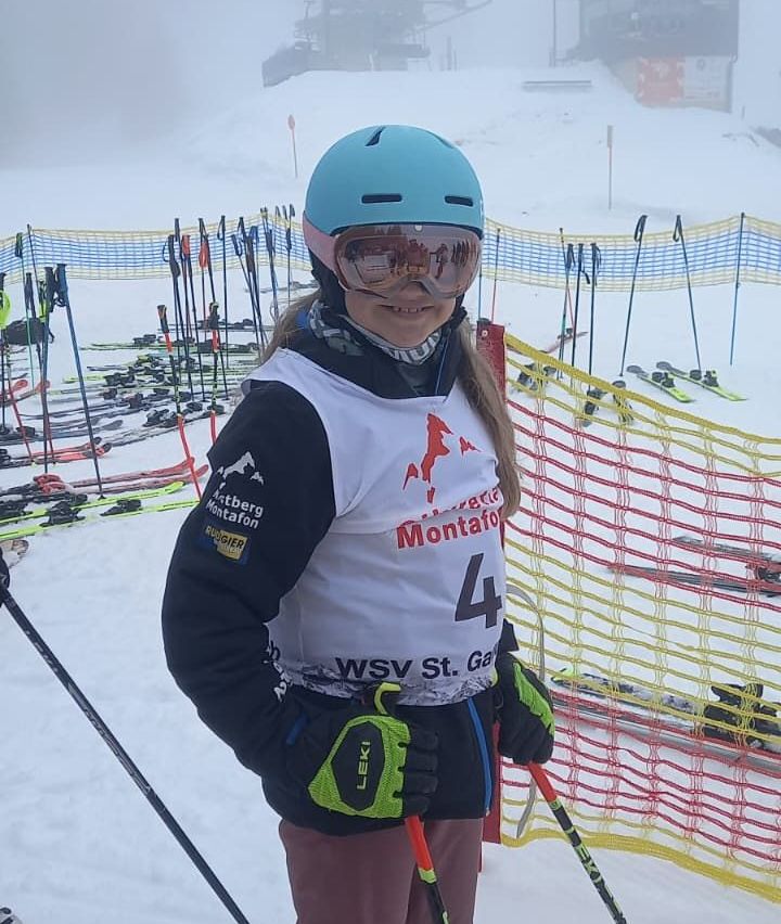 Eine junge Skifahrerin mit der Nummer 4 auf ihrem Trikot, mit Helm, Brille und Skihandschuhen, steht bei schneebedecktem Wetter mit Skiern und Stöcken im Hintergrund.