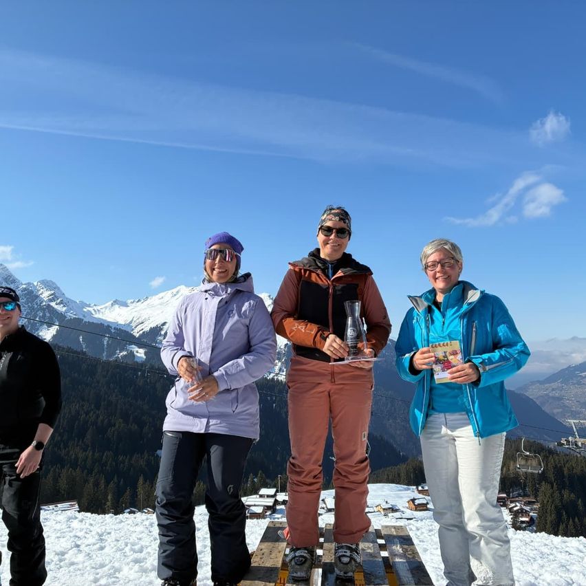 Vier Personen stehen auf einem verschneiten Berg, zwei Frauen halten Trophäen, eine mit einem Buch. Dahinter Berge, die mit Schnee bedeckt sind.