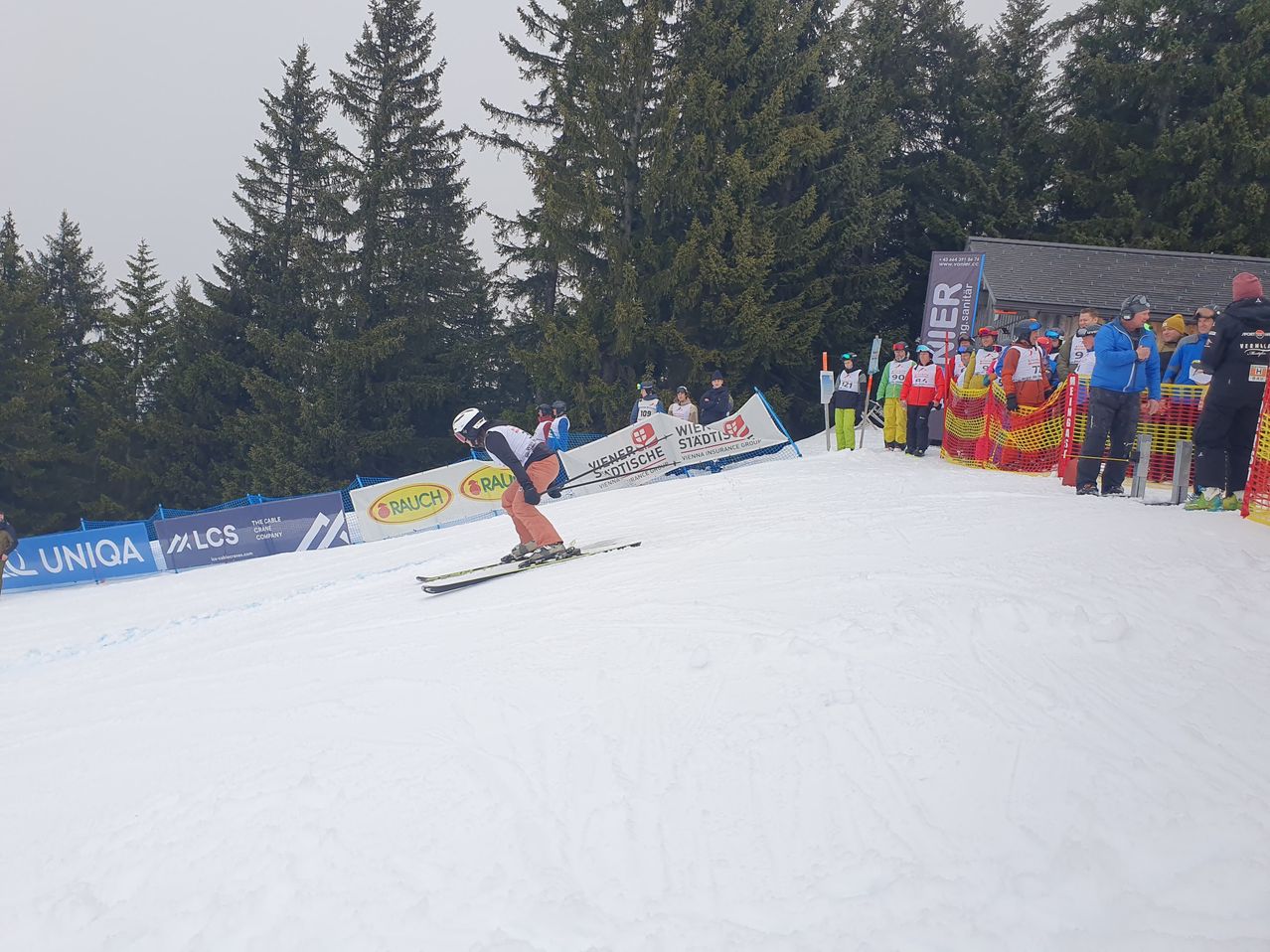 Ein Skifahrer fährt einen schneebedeckten Hang hinunter, mit Bäumen und Menschen im Hintergrund. Mehrere Personen stehen hinter Barrieren und beobachten den Skifahrer.
