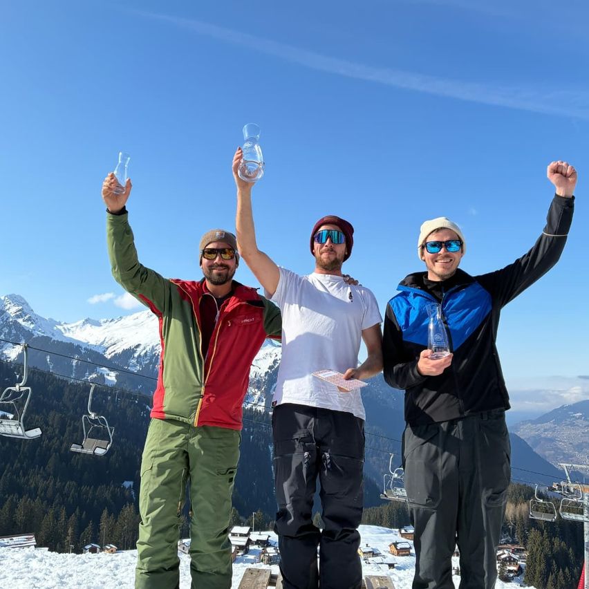 Drei Männer stehen auf einem verschneiten Berg, halten klare Glastrophäen und feiern. Sie tragen Winterkleidung und sind von Skiliften und Bäumen umgeben.