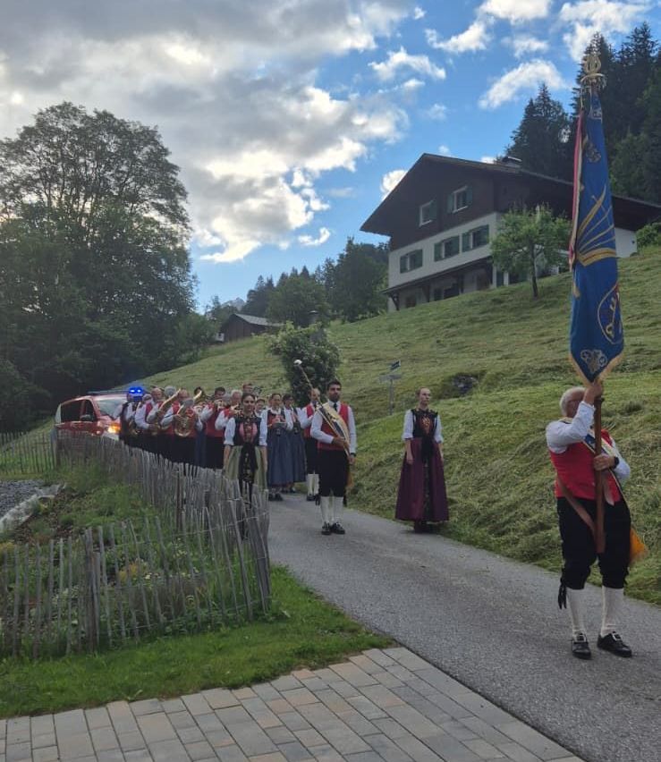Ein Marschkapelle spielt Instrumente auf einem Hügel mit einer Fahne vorne, ein Haus im Hintergrund.