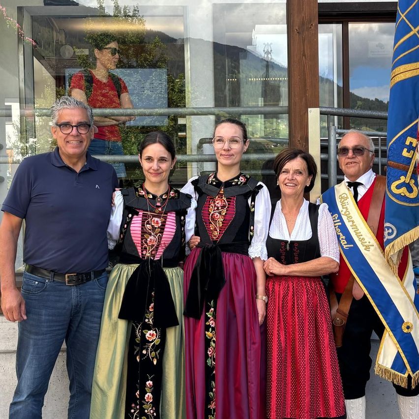 Fünf Personen, darunter zwei Männer und drei Frauen, stehen gemeinsam für ein Foto. Die Frauen tragen traditionelle Kostüme und die Männer sind lässig gekleidet. Das Foto wurde vor einem Gebäude mit einer Glaswand aufgenommen.