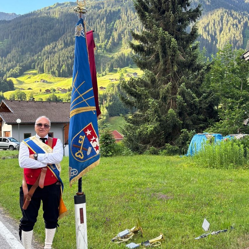 Ein Mann steht neben einem Fahnenmast, lächelt und trägt ein traditionelles Outfit. Dahinter befindet sich ein Rasenfeld mit verstreuten Musikinstrumenten. Im Hintergrund zeigt sich eine malerische Berglandschaft mit Häusern und Bäumen.