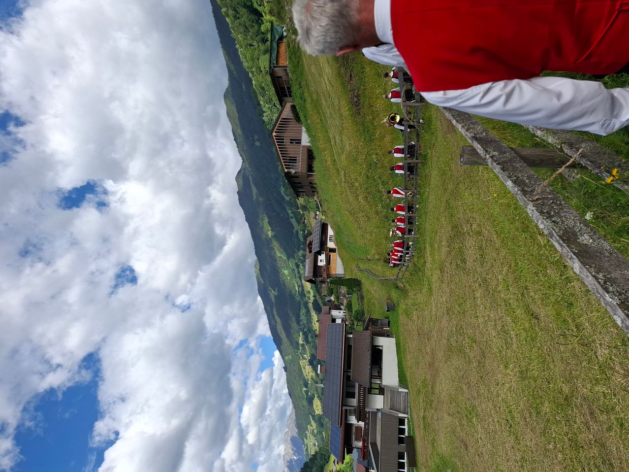 Luftaufnahme eines Tals mit Häusern, einem grasigen Feld und Musikern in roten und weißen Kostümen. Der Himmel ist bewölkt.