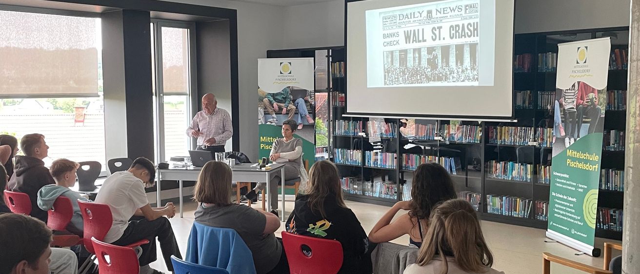 Eine Präsentation über den Wall Street Crash wird auf einer Projektionsleinwand in einer Bibliothek gezeigt. Ein Mann steht vor der Leinwand, während eine Gruppe von Menschen auf Stühlen davor sitzen.