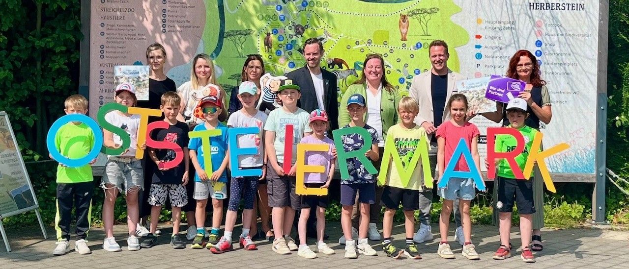 Eine Gruppe von Erwachsenen und Kindern steht vor einem Wandbild und hält Buchstaben hoch, die 'STEINERMARK' buchstabieren. Sie lächeln und scheinen für ein Foto zu posieren. Dahinter ist eine Wand mit einer Karte zu sehen.