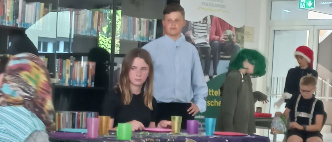 Mehrere Personen sitzen um einen Tisch in einer Bibliothek; ein Mädchen in Schwarz und ein Junge in Blau sitzen am Tisch mit Tassen und Tellern. Dahinter steht eine Person mit grünen Haaren.