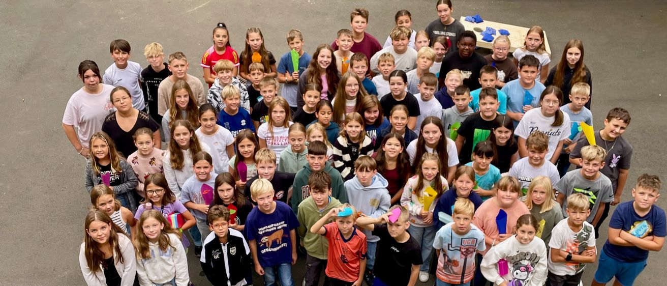 Eine Gruppe von Kindern und Erwachsenen steht in einem großen Kreis und hält Gegenstände in der Hand. Sie sind in einem Außenbereich, mit einem Gebäude und einem grünen Rasen im Hintergrund.