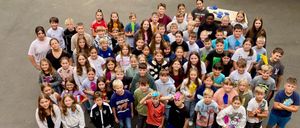 Eine Gruppe von Kindern und Erwachsenen steht in einem großen Kreis und hält Gegenstände in der Hand. Sie sind in einem Außenbereich, mit einem Gebäude und einem grünen Rasen im Hintergrund.