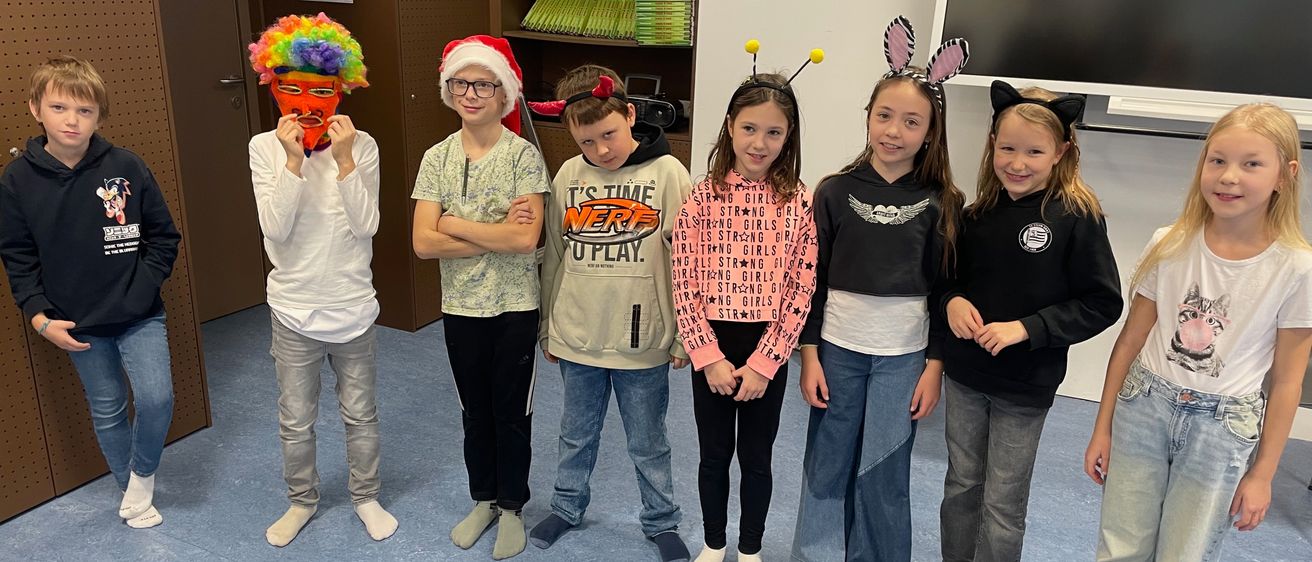 Eine Gruppe von Kindern in verschiedenen Kostümen steht zusammen. Einer trägt eine Maske, ein anderer einen Weihnachtsmannhut und andere verschiedene Accessoires. Sie stehen in einem Klassenzimmer mit Bücherregalen und einem Monitor im Hintergrund.