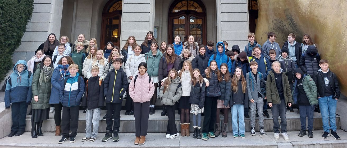 Eine Gruppe von Schülern steht auf den Stufen eines großen Gebäudes. Das Gebäude zeigt eine goldene Skulptur, einen Balkon mit Geländer und ein Schild mit Text. Die Schüler sind warm gekleidet, einige tragen Rucksäcke.
