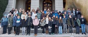 Eine Gruppe von Schülern steht auf den Stufen eines großen Gebäudes. Das Gebäude zeigt eine goldene Skulptur, einen Balkon mit Geländer und ein Schild mit Text. Die Schüler sind warm gekleidet, einige tragen Rucksäcke.