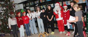 Eine Gruppe von Kindern und zwei Erwachsenen, die als Weihnachtsmann verkleidet sind, stehen in einer Bibliothek. Sie halten Mikrofone in der Hand und scheinen sich auf eine Aufführung vorzubereiten.