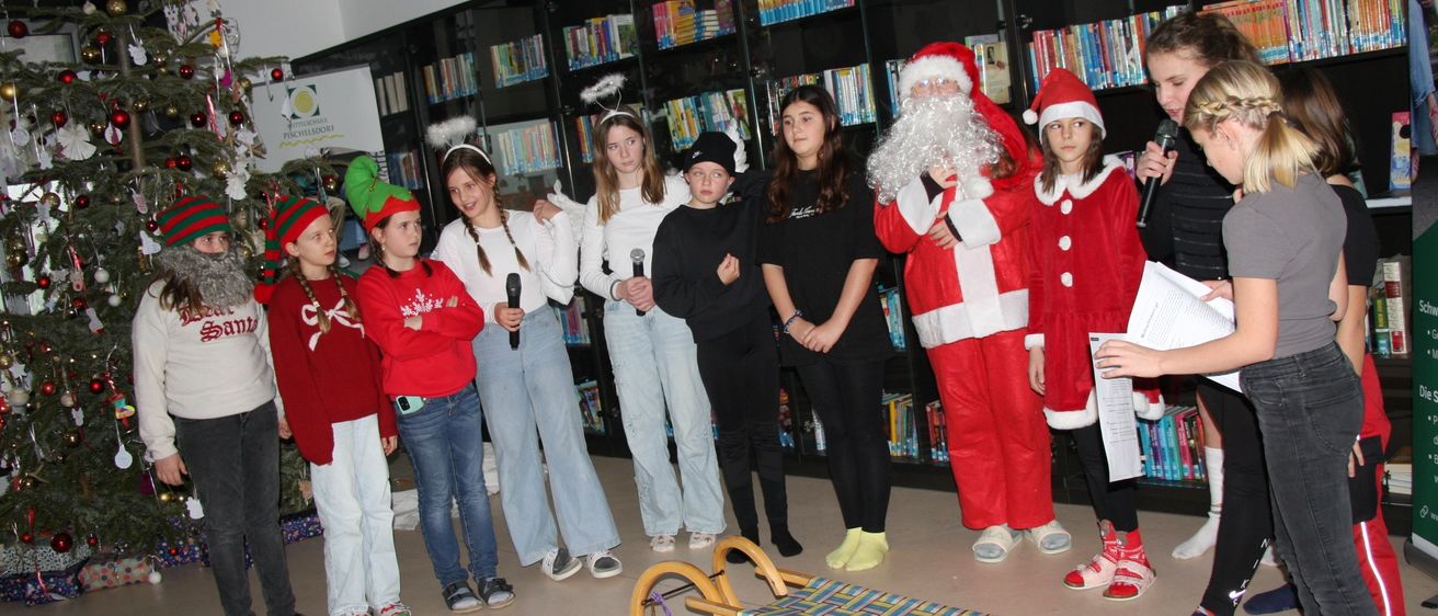Eine Gruppe von Kindern und zwei Erwachsenen, die als Weihnachtsmann verkleidet sind, stehen in einer Bibliothek. Sie halten Mikrofone in der Hand und scheinen sich auf eine Aufführung vorzubereiten.