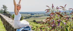 Eine Frau sitzt auf einer Steinmauer, die Arme zum Jubel erhoben, in einem weißen Kleid. Sie blickt auf eine ländliche Landschaft mit viel Grün und entfernten Hügeln.