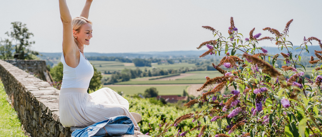 Eine Frau sitzt auf einer Steinmauer, die Arme zum Jubel erhoben, in einem weißen Kleid. Sie blickt auf eine ländliche Landschaft mit viel Grün und entfernten Hügeln.