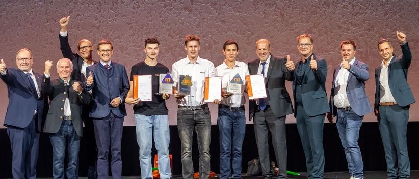 Eine Gruppe von Menschen in Anzügen und Krawatten, die auf einer Bühne stehen, Urkunden halten und eine Daumen-hoch-Geste machen.