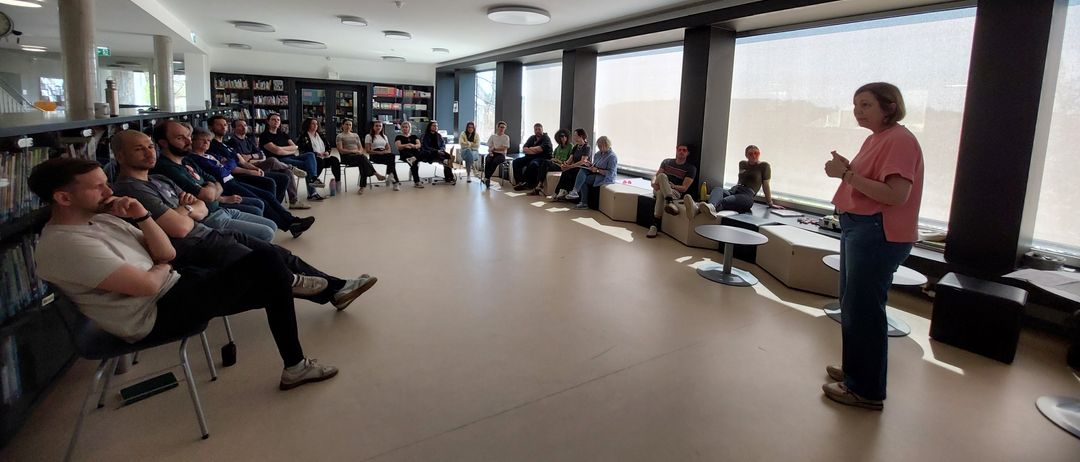 Eine Frau steht an einem Podium in einem Klassenzimmer. Eine Gruppe von Menschen sitzt im Kreis um sie herum. Einige Menschen stehen in der Nähe von Fenstern.