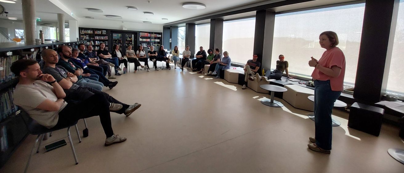 Eine Frau steht an einem Podium in einem Klassenzimmer. Eine Gruppe von Menschen sitzt im Kreis um sie herum. Einige Menschen stehen in der Nähe von Fenstern.