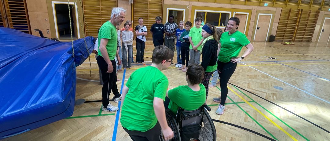 Eine Sporthalle, in der sich mehrere Personen in grünen T-Shirts, einige stehend und einige in Rollstühlen, um eine Frau versammeln und sich unterhalten. An der Wand im Hintergrund ist ein Basketballkorb befestigt.