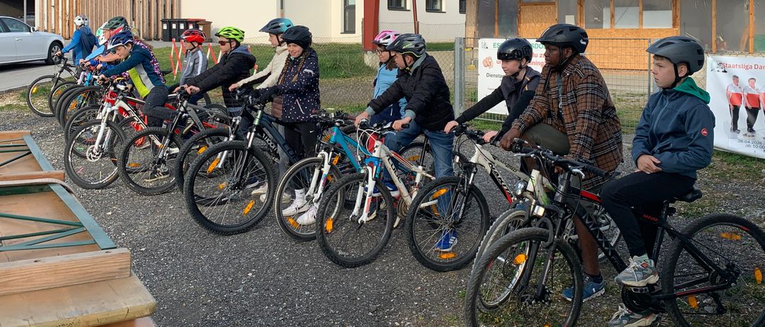 Eine Gruppe junger Leute mit Helmen steht mit ihren Fahrrädern in einem Parkplatz. Dahinter befinden sich einige Gebäude mit roten Dächern.
