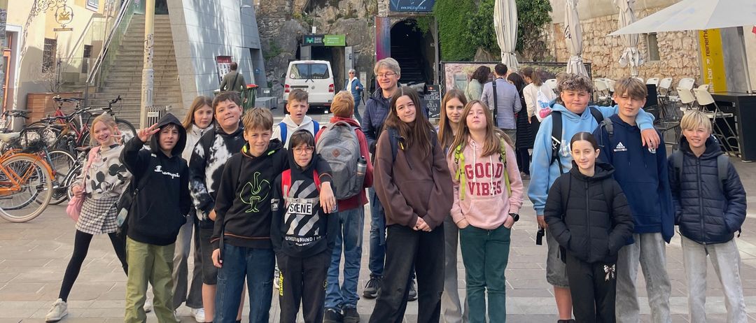Eine Gruppe von Kindern und Erwachsenen steht draußen vor einem Gebäude mit einer Treppe, die zu einer Klippe führt. In der Nähe sind Fahrräder geparkt und im Hintergrund befindet sich ein Banner.