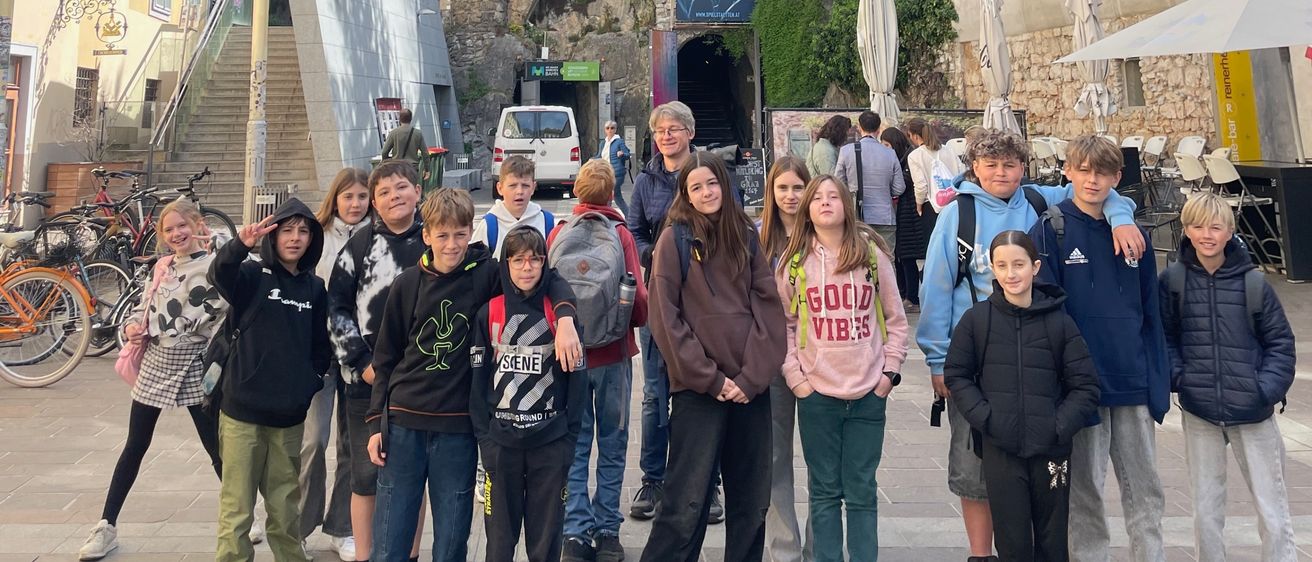 Eine Gruppe von Kindern und Erwachsenen steht draußen vor einem Gebäude mit einer Treppe, die zu einer Klippe führt. In der Nähe sind Fahrräder geparkt und im Hintergrund befindet sich ein Banner.