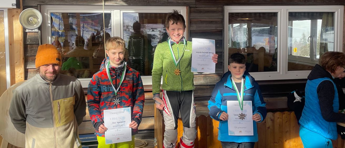 Vier Jungen mit Medaillen stehen auf einem Podest vor einer Holzkonstruktion. Sie tragen Winterkleidung und halten Urkunden in den Händen. Der Junge in der Mitte trägt rote Skischuhe. Dahinter steht eine Person in der Nähe eines Tisches.