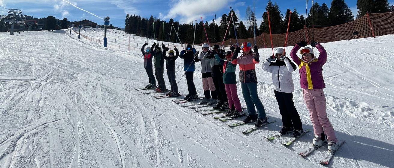 Eine Gruppe von Menschen in Skiausrüstung steht auf einem verschneiten Hang und hält ihre Skistöcke unter einem blauen Himmel mit verstreuten Wolken.