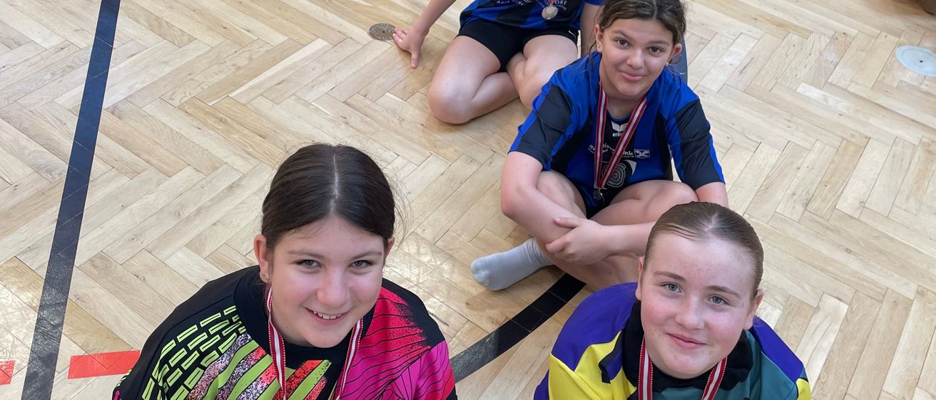 Eine Gruppe junger Mädchen mit Medaillen um den Hals sitzt auf dem Boden in einer Turnhalle. Sie alle lächeln und posieren für ein Foto. Der Boden hat Markierungen und einen weißen Kreis.