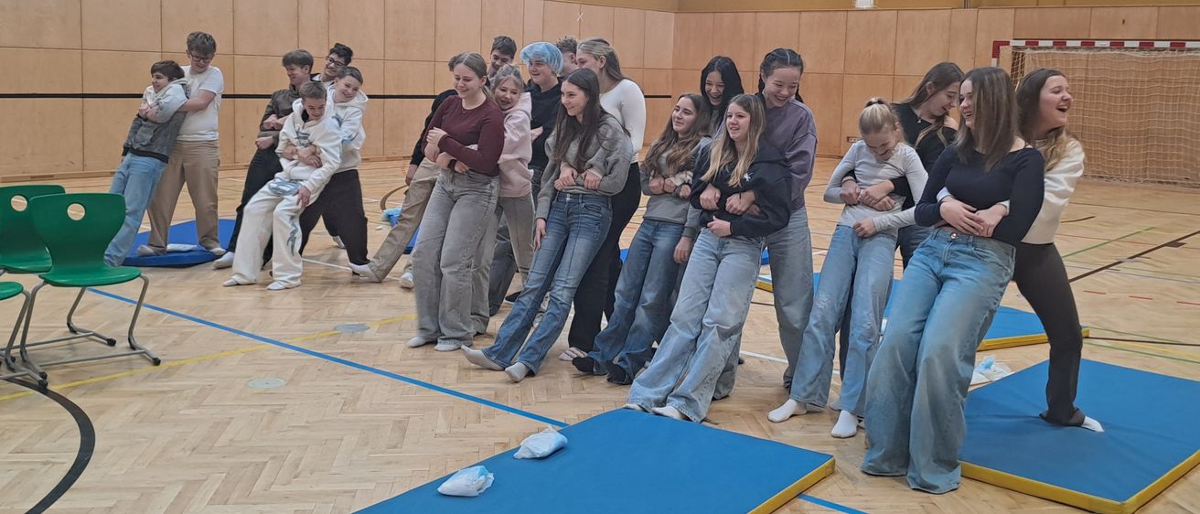 Eine Gruppe von Menschen steht in einer Turnhalle, einige auf blauen Matten, lächelt und posiert für ein Foto. Ein Basketballkorb und ein grüner Stuhl sind im Hintergrund zu sehen.