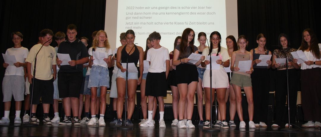 Eine Gruppe von Schülern auf der Bühne, die Papiere halten, wahrscheinlich bei einer Schulveranstaltung. Ein Projektionsbildschirm hinter ihnen zeigt Text.