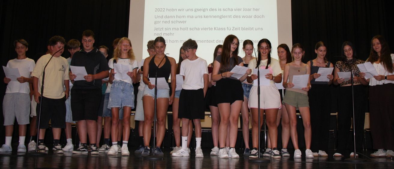 Eine Gruppe von Schülern auf der Bühne, die Papiere halten, wahrscheinlich bei einer Schulveranstaltung. Ein Projektionsbildschirm hinter ihnen zeigt Text.