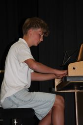 Ein junger Junge in einem weißen T-Shirt und Jeansshorts spielt konzentriert das Klavier, vor einem schwarzen Vorhang sitzend.