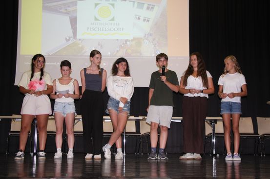 Eine Gruppe von Schülern steht auf der Bühne, hält Papiere und ein Mikrofon. Ein Projektionsbildschirm hinter ihnen zeigt ein Schulgebäude.