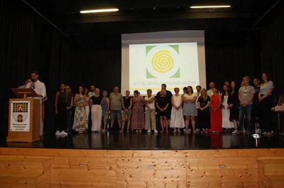 Eine Gruppe von Menschen steht auf der Bühne und sieht das Publikum an, während eine große Leinwand im Hintergrund das Logo der Mittelschule Pischelsdorf zeigt.