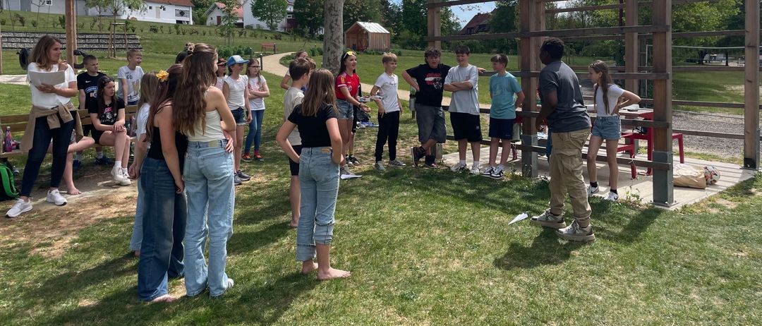 Eine Gruppe von Schülern versammelt sich in einem Park. Einige stehen in der Nähe einer Holzstruktur, während andere in lässiger Kleidung herumstehen. Die Umgebung ist im Freien, mit einem klaren blauen Himmel im Hintergrund.