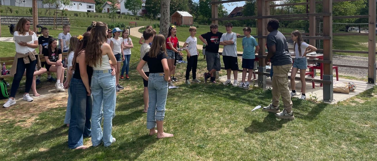 Eine Gruppe von Schülern versammelt sich in einem Park. Einige stehen in der Nähe einer Holzstruktur, während andere in lässiger Kleidung herumstehen. Die Umgebung ist im Freien, mit einem klaren blauen Himmel im Hintergrund.