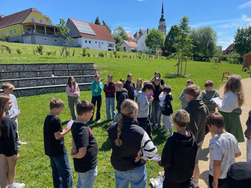 Eine Gruppe von Kindern und Jugendlichen steht im Kreis auf einem Rasenfeld mit einer Kirche und Häusern im Hintergrund.