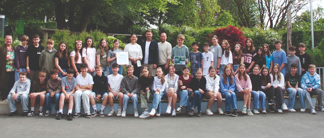 Eine Gruppe von Schülern und Lehrern posiert für ein Foto im Park. Zwei Personen halten Urkunden. Bäume und Pflanzen umgeben das Gebiet.