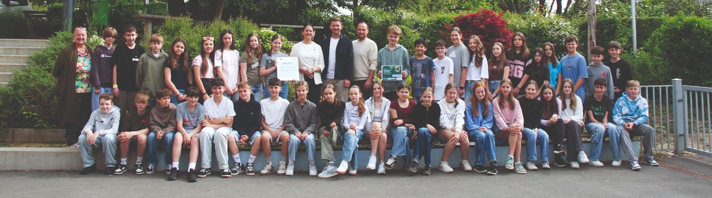 Eine Gruppe von Schülern und zwei Erwachsene stehen zusammen und posieren für ein Foto vor grünen Pflanzen.