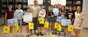 Eine Gruppe junger Kinder und ein erwachsener Mann posieren für ein Foto in einer Bibliothek. Sie halten gelbe Taschen und Zeichnungen. Der Hintergrund ist ein Banner mit einem Logo.