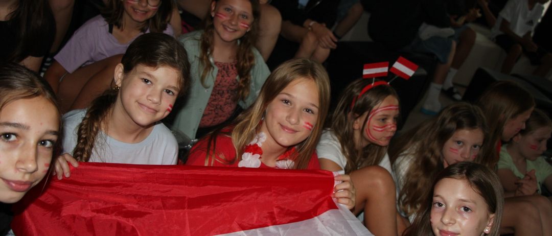 Gruppe von Kindern mit österreichischen Flaggen im Gesicht gemalt, die eine große österreichische Flagge halten.