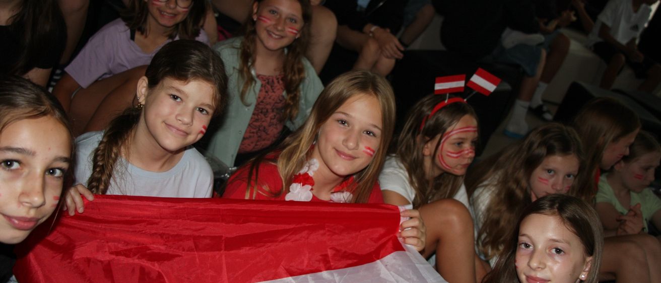 Gruppe von Kindern mit österreichischen Flaggen im Gesicht gemalt, die eine große österreichische Flagge halten.