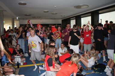 Eine Gruppe junger Leute versammelt sich drinnen, einige stehen und andere sitzen, wobei mehrere Menschen die Hände heben. Sie tragen T-Shirts in verschiedenen Farben und einige tragen Hüte. Sie scheinen ein Ereignis zu feiern oder daran teilzunehmen.