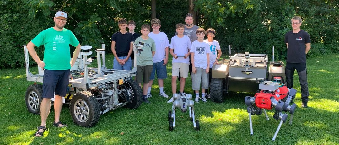 Eine Gruppe von Menschen, darunter Erwachsene und Kinder, steht in einem Park mit vier Robotern, von denen einer ein großer Rover ist.