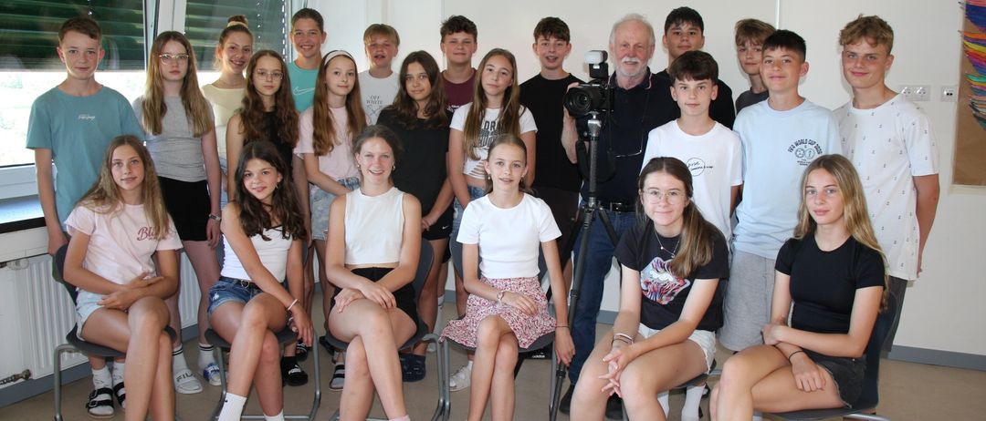 Eine Gruppe junger Menschen und ein Mann versammeln sich in einem Raum, möglicherweise für ein Fotoshooting. Sie sitzen auf Stühlen, ein Mann steht hinter einer Kamera mit Stativ. Der Raum hat weiße Wände und Fenster mit Jalousien.